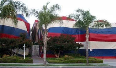 A home covered in a large, striped fumigation tent with a real estate sign in front