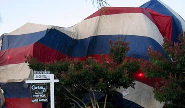 A home covered in a large, striped fumigation tent with a real estate sign in front