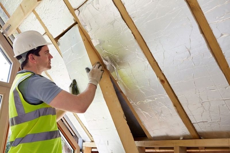 A man in a safety vest and hardhat installing a new piece of insulation into the underside of a home's roof