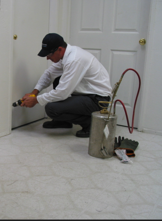 A man in a white, collared shirt and blue baseball cap spraying pesticide around the edge of a doorway